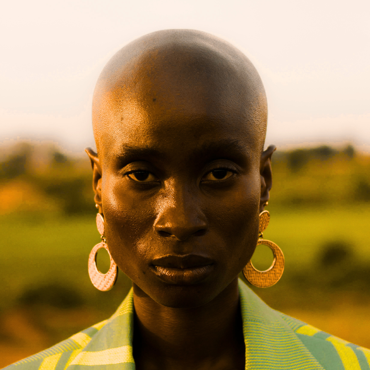 A beautiful, bald, black woman with a fierce look of strength and empowerment wearing large, gold, drop earrings and a green and yellow blazer.